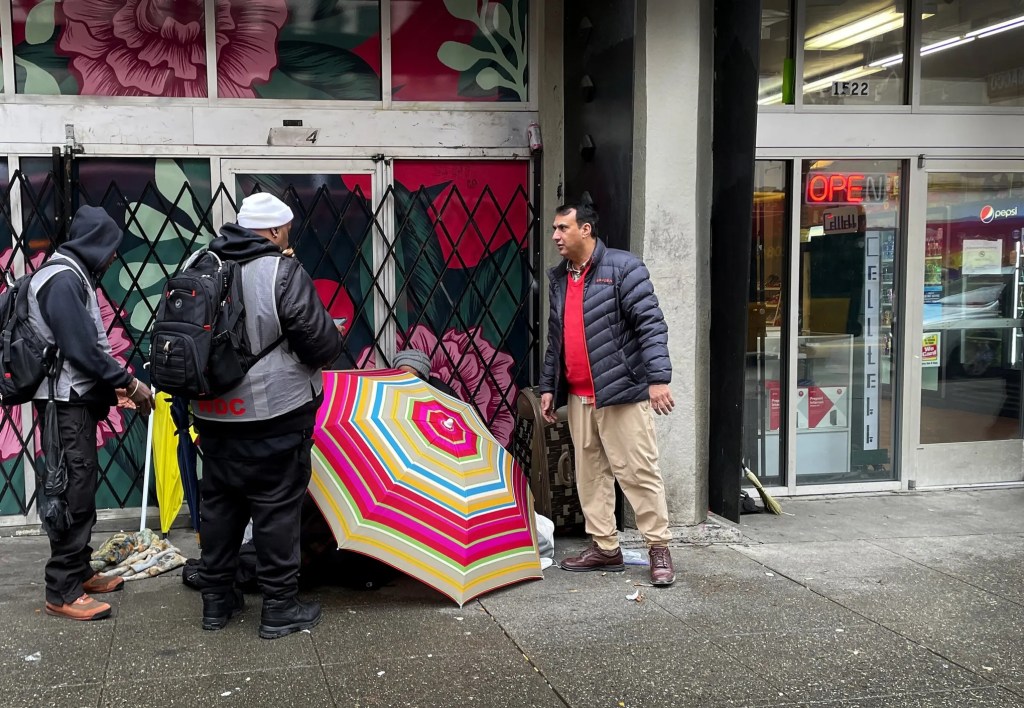 We Deliver Care team members, left, talk to a woman yelling from behind an umbrella, while Maqsood Choudhary, right, co-owner of International Cigar and Tobacco next door, monitors the situation on Third Avenue between Pine and Pike streets on Monday. Choudhary said We Deliver Care did diffuse the situation but feels nothing has changed here since the pandemic. (Ken Lambert / The Seattle Times)